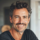 Homme souriant avec une barbe, fond intérieur lumineux.