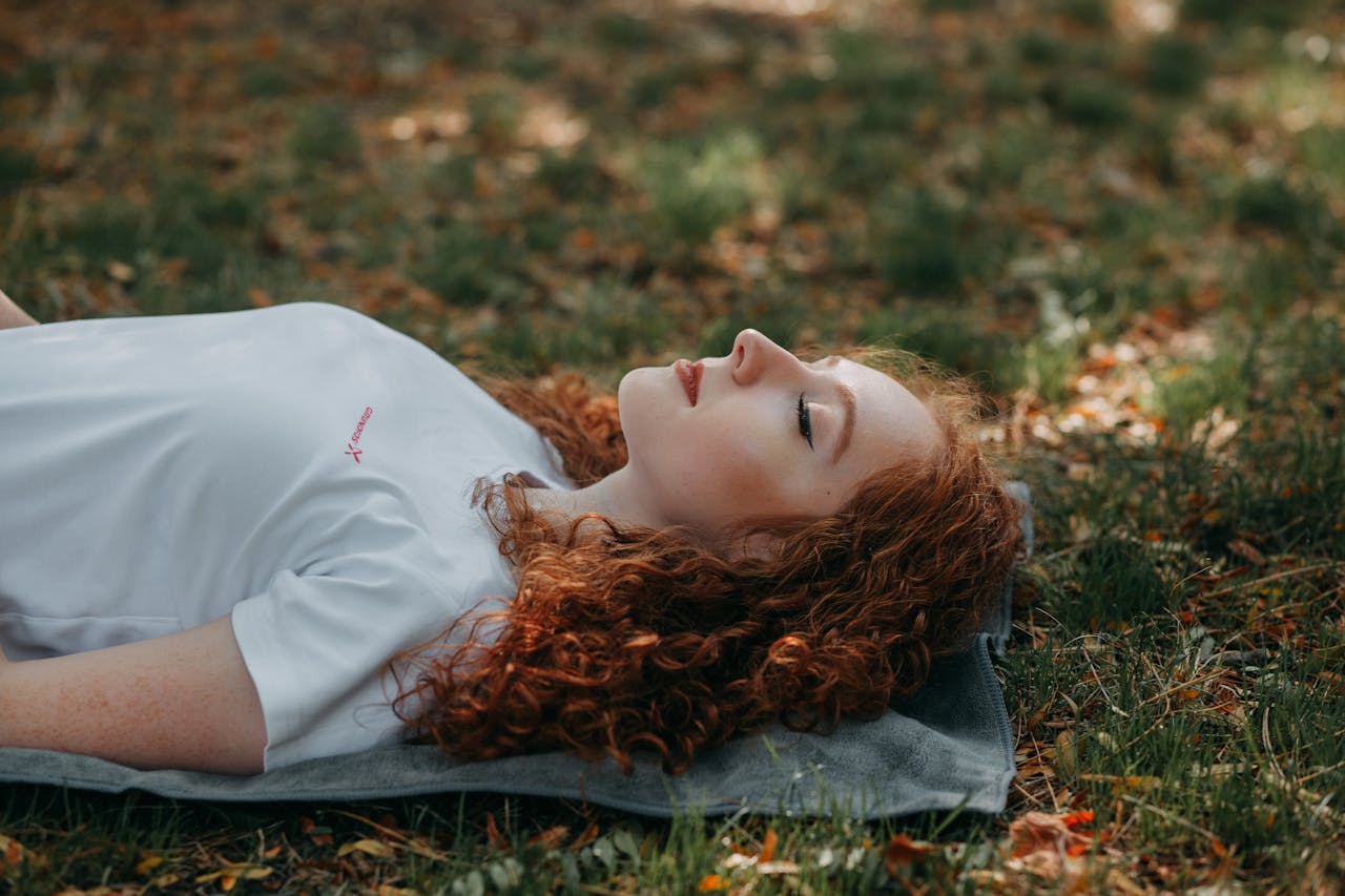 about-us-img Adult woman with red hair resting peacefully outdoors on a sunny day, surrounded by nature.
